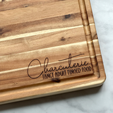 Wooden cutting board with 'Charcuterie Fancy Adult Finger Food' engraving on a marble surface